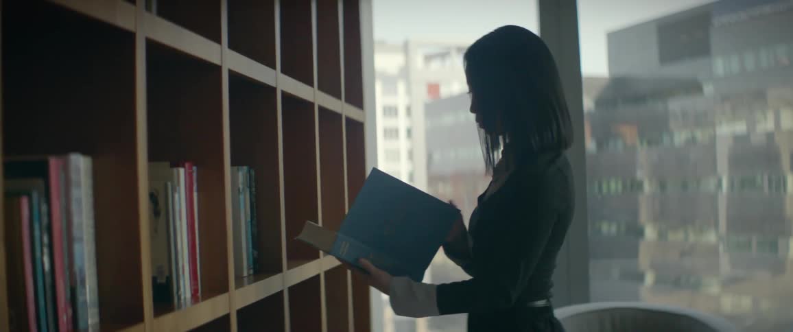 A professional woman walks up to a large bookshelf in a modern office, selects a book, and walks away with it