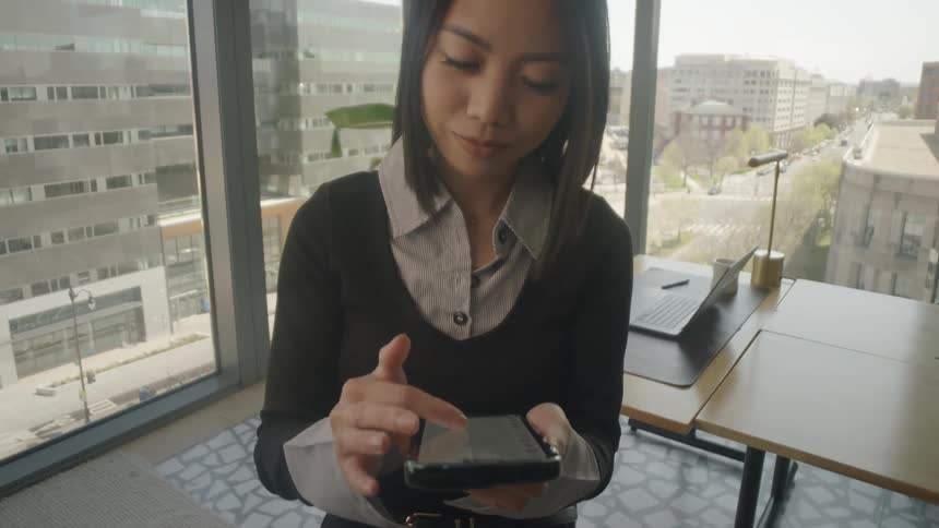 Dolly out shot of an Asian businesswoman scrolling on her smartphone in a modern high-rise office with a city view