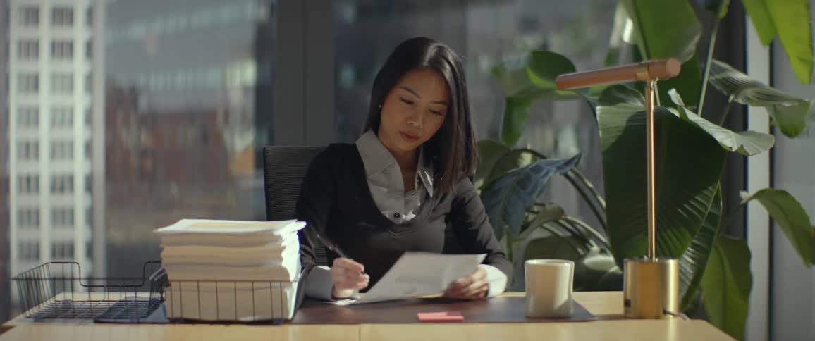 A young Asian businesswoman signs a document at her desk in a sunlit modern office, then looks up and smiles confidently at the camera