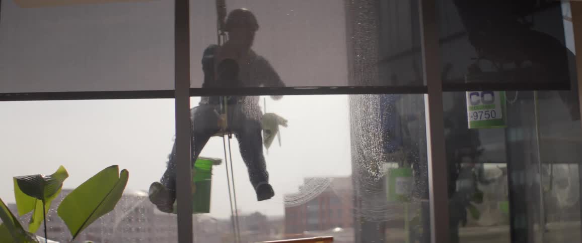 A window washer suspended in a harness cleans the exterior glass of a skyscraper in slow motion, as seen from inside the building