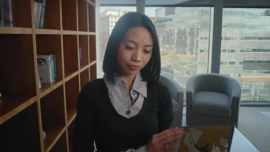 A young Asian woman selects a large book from a wooden bookshelf in a modern office library overlooking the city
