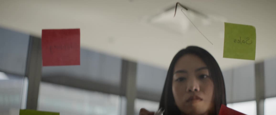 A focused Asian businesswoman strategizes and plans a project by writing on a glass wall with sticky notes in a modern office