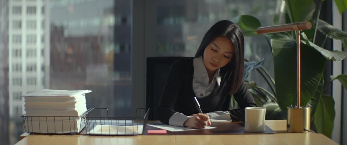 An Asian businesswoman at her desk in a sunlit high-rise office signs a college acceptance letter and then pauses to look thoughtfully out the window