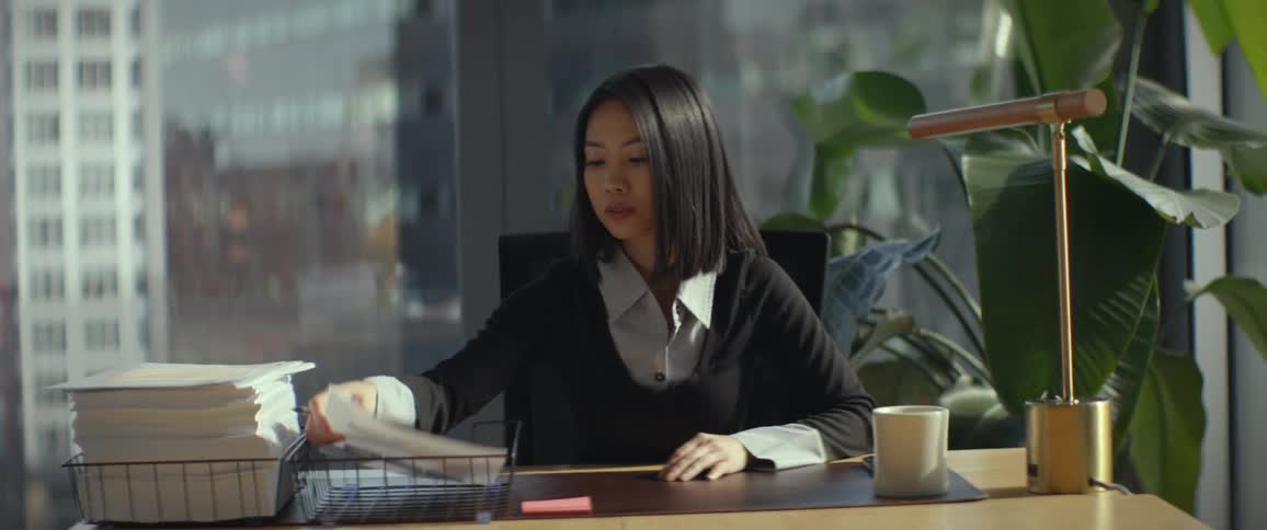 A young Asian professional meticulously reviews and signs important documents while working at her desk in a modern, sunlit high-rise office