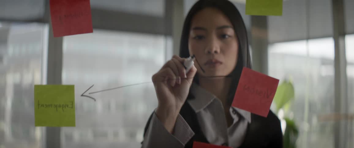 A focused Asian businesswoman brainstorms a project strategy using colorful sticky notes and a marker on a glass wall in a modern office
