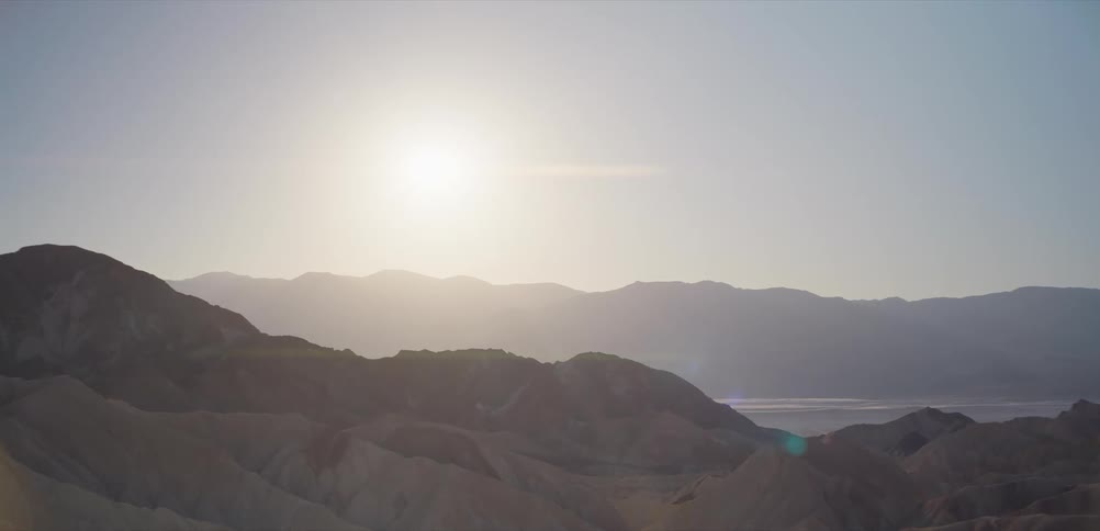A bright sun hangs over the vast, hazy desert mountain range at sunrise, casting a lens flare across the landscape