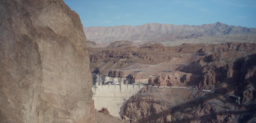 A wide, static shot of the iconic Hoover Dam set against a backdrop of rugged mountains and canyons on a clear day