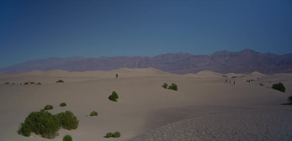 Hikers traverse the expansive, rolling sand dunes of a vast desert with mountains on the horizon