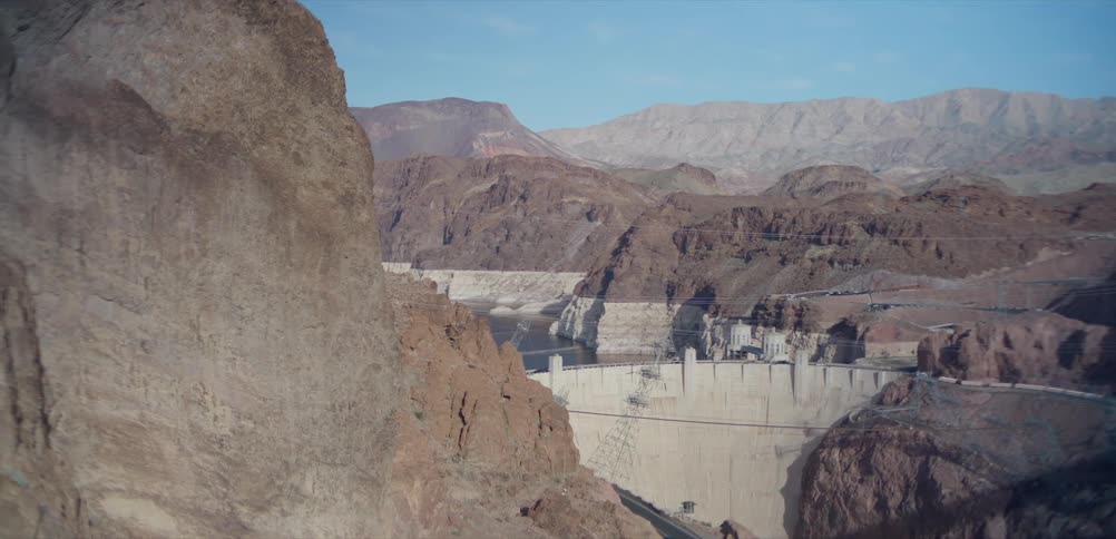 A high-angle, wide shot captures the immense Hoover Dam and Lake Mead set within the rugged, rocky canyon landscape