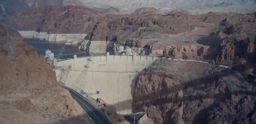 A high-angle tilt shot reveals the Hoover Dam and Lake Mead on the Colorado River in a vast desert canyon