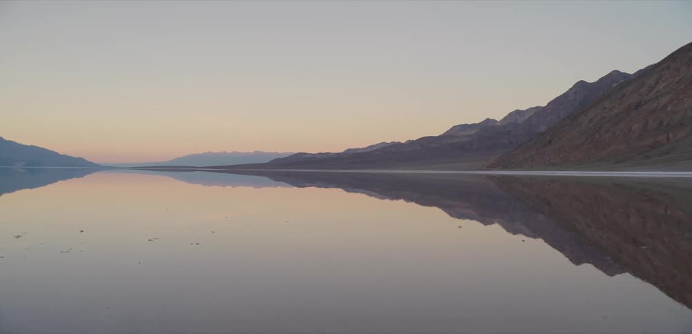 A tranquil desert landscape features mountains perfectly mirrored in the still, glassy water at dawn