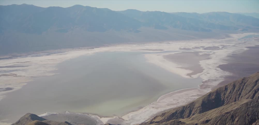 A stunning high-angle, wide shot captures the immense scale of the Badwater Basin salt flats nestled within the hazy mountains of Death Valley