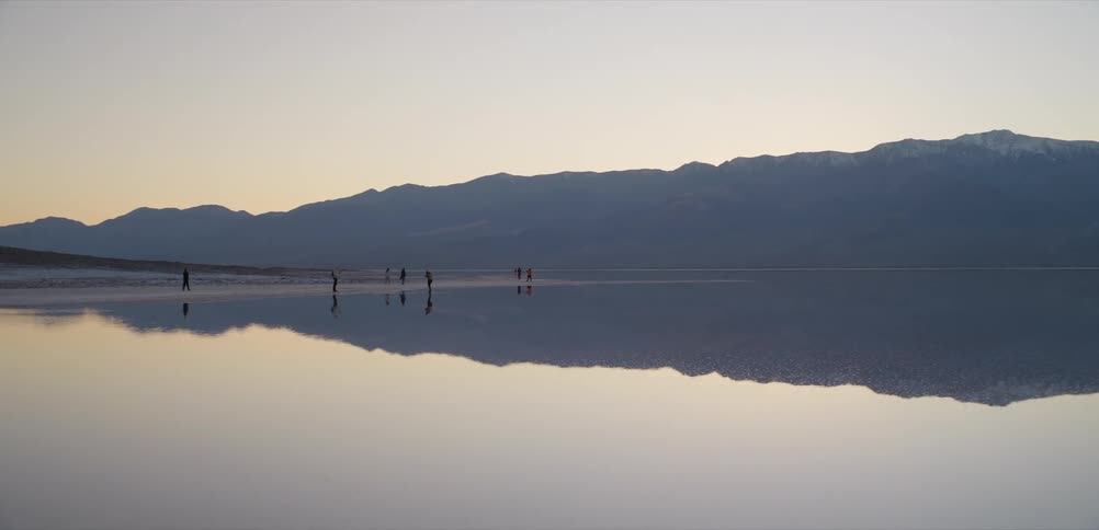 A group of people walk on a reflective lake at dusk, with their silhouettes and the mountains perfectly mirrored on the calm water's surface