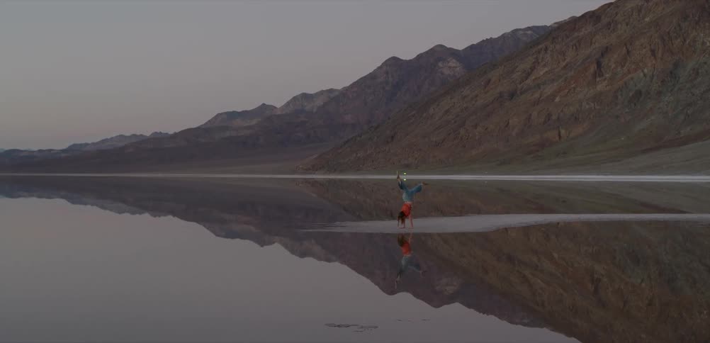 A woman holds a handstand on the edge of a serene, reflective lake, creating a perfect mirror image with the vast mountain range behind her
