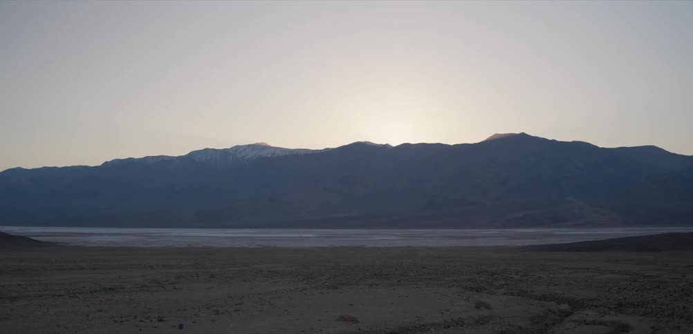 A serene sunrise glows from behind a snow-capped mountain range in a vast desert landscape