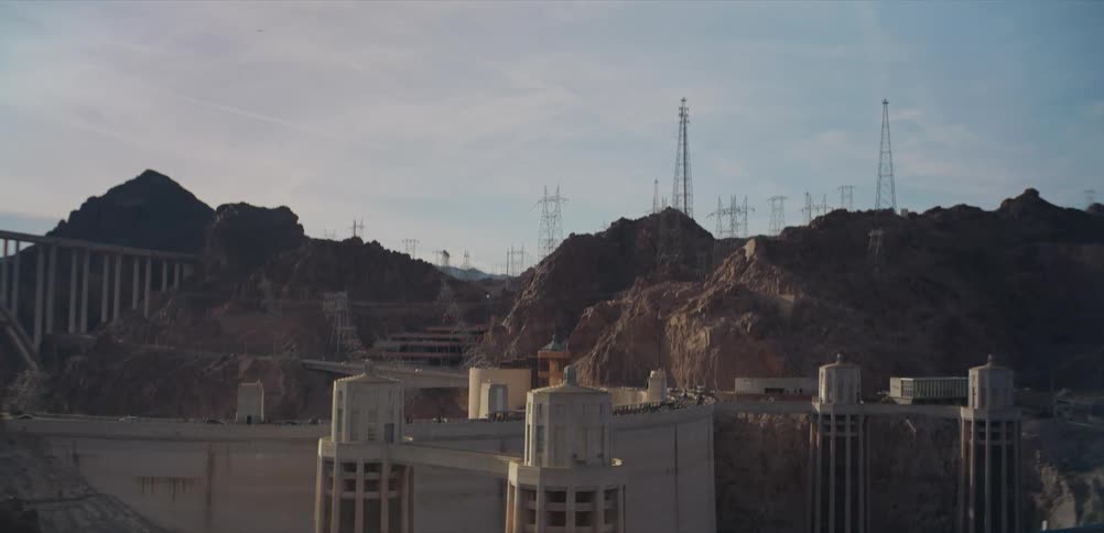 A static wide shot of the Hoover Dam showcases the massive structure and surrounding power lines set against a rugged mountain landscape