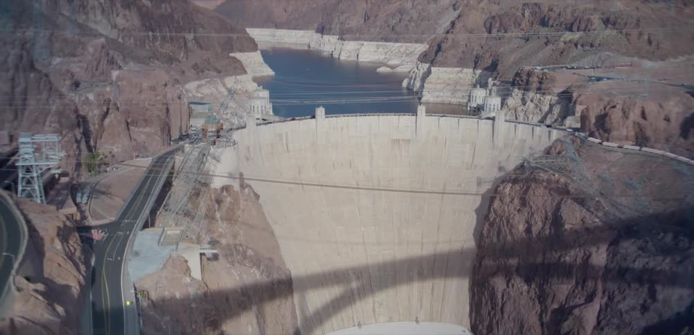 A slow upward tilt reveals the massive Hoover Dam, its powerhouse, and the low water levels of Lake Mead in the surrounding canyon
