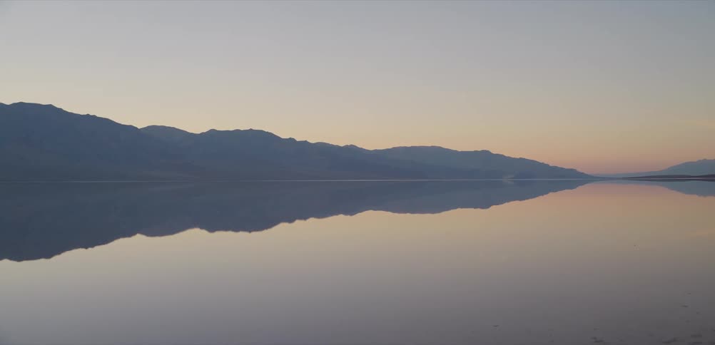 Distant mountains cast a perfect reflection on the calm, glassy surface of a lake during a tranquil sunrise