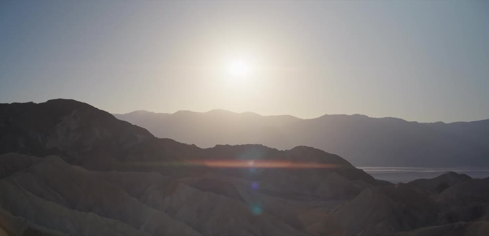 The bright sun creates a lens flare over a vast, hazy desert mountain landscape