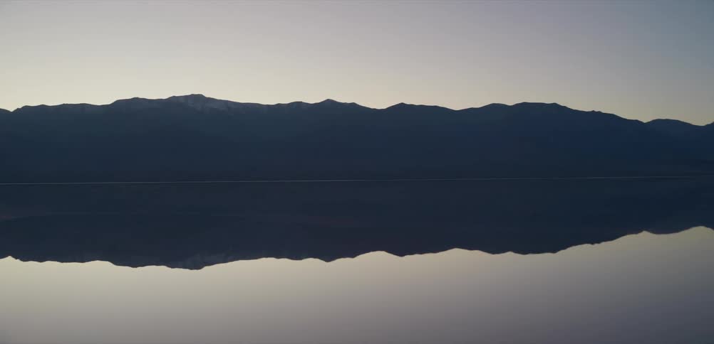 A serene panoramic shot reveals the majestic mountains of Death Valley reflected perfectly in the still waters of the Badwater Basin salt flats at dusk