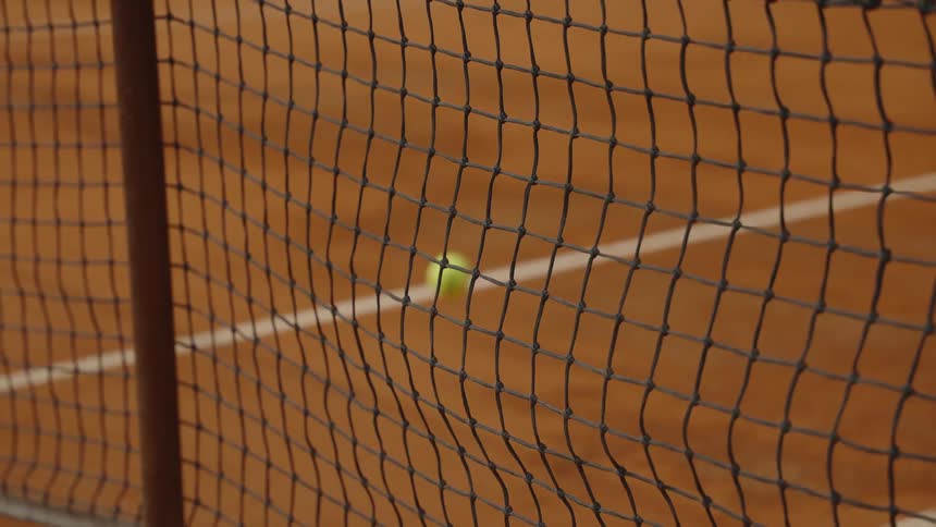 A tennis ball bounces in slow motion on a clay court with the net in the foreground
