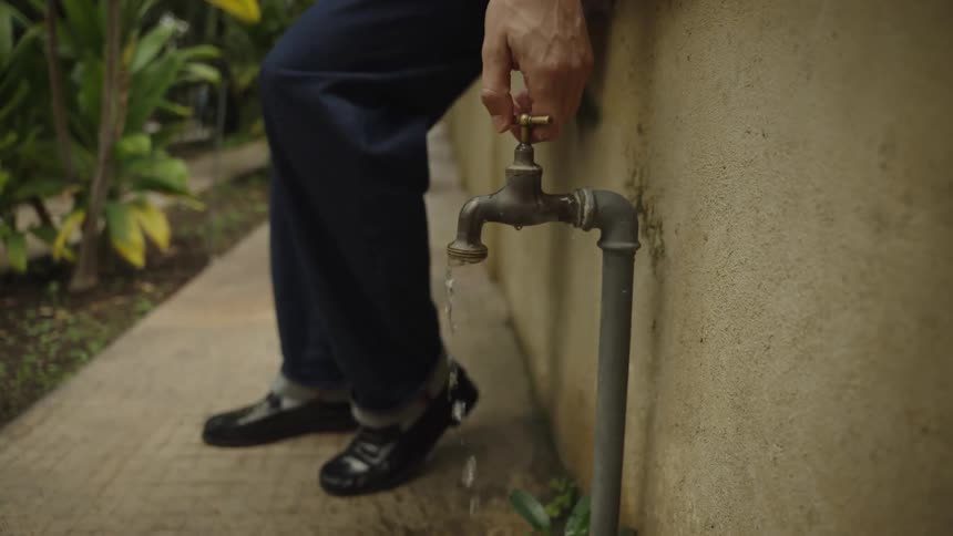 A man turns off a weathered outdoor water faucet, with the water flow stopping in slow motion to conserve resources