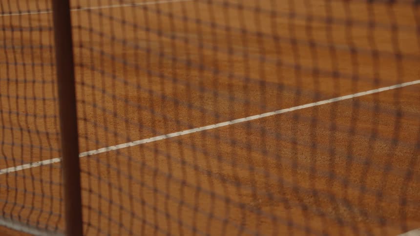 A tennis ball bounces in slow motion along the white line on a clay court, viewed through the net