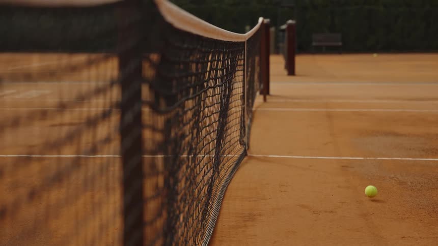 A tennis ball hits the net in slow motion on an empty clay court