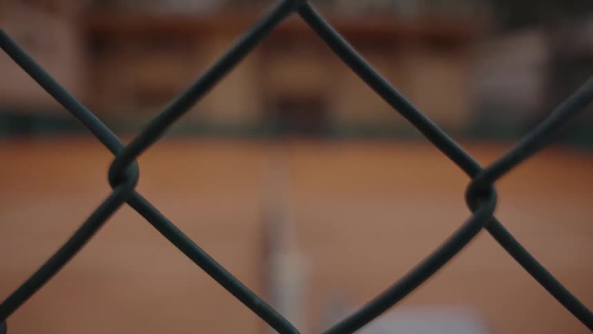 A rack focus shot reveals an empty clay tennis court through a chain-link fence