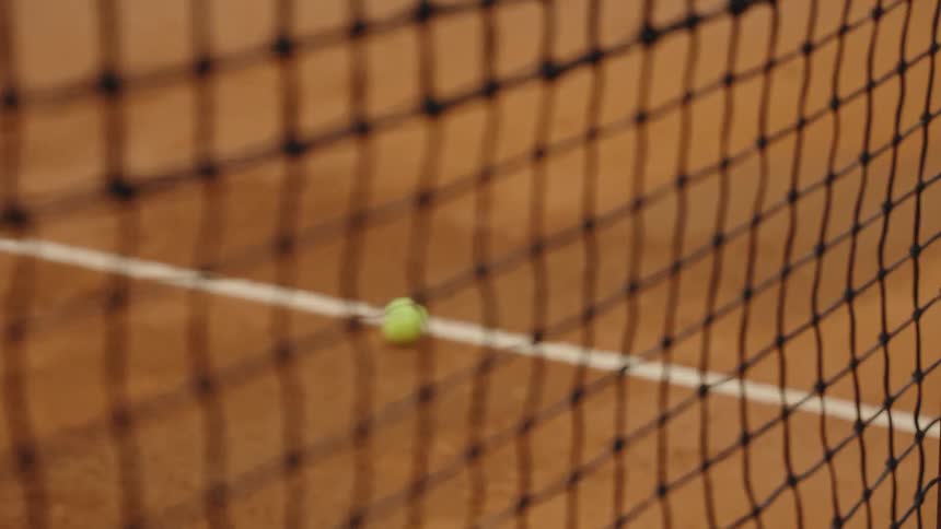 Close-up of a tennis net with a rack focus to a yellow tennis ball rolling to a stop on the white line of a clay court