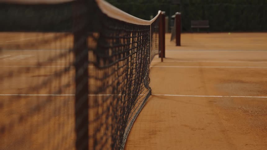 A tennis ball flies over the net in slow motion on an empty clay court at sunset