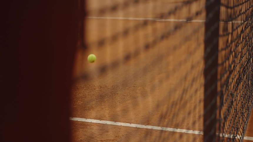 A tennis ball bounces in slow motion across a sunlit clay court as seen through the net