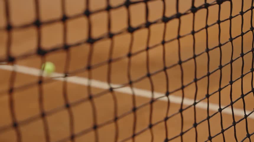 A close-up shot of a tennis net with a focus pull to a yellow tennis ball on a clay court