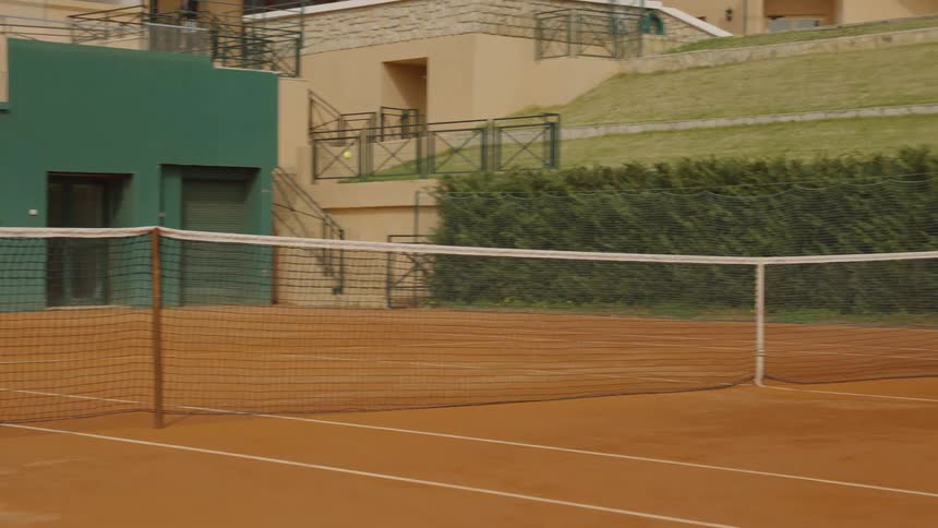 A tennis ball is hit in slow motion and flies over the net on an empty clay court