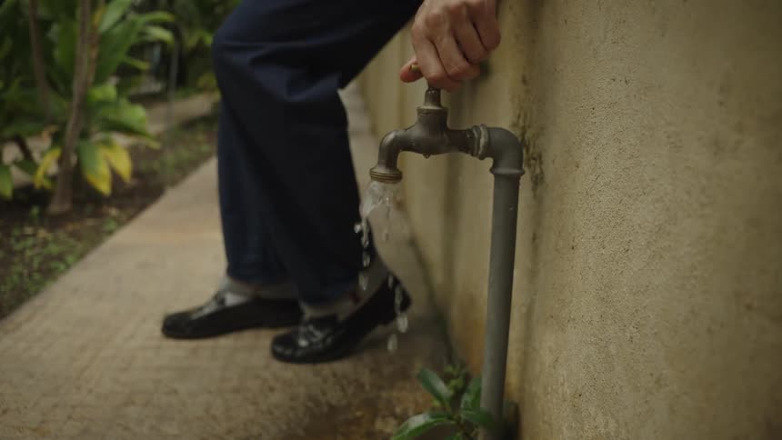 A person's hand turns the handle of an outdoor water faucet, releasing a stream of water in slow motion