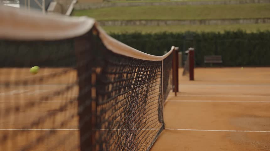 A yellow tennis ball bounces in slow motion on a clay court, seen from a low angle next to the net