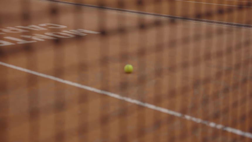 Slow focus reveals a lone tennis ball resting on a clay court through a tennis net