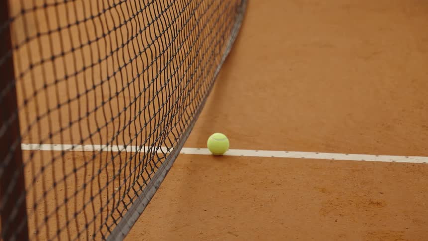 A close-up view of a bright yellow tennis ball resting on the white line beside the net on a clay court