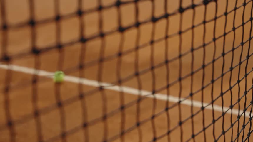 Slow motion close-up of a tennis ball rolling onto the white line of a clay court, viewed through the net with a rack focus