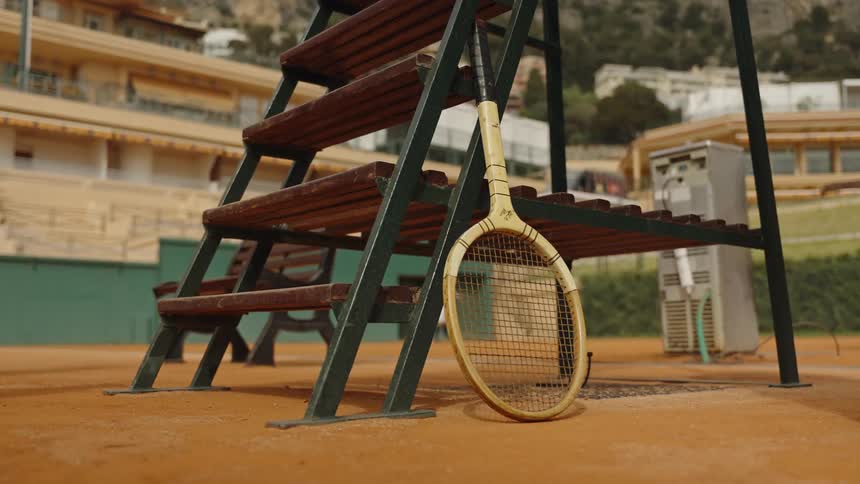 A vintage wooden tennis racket rests against an umpire's chair on a classic clay court