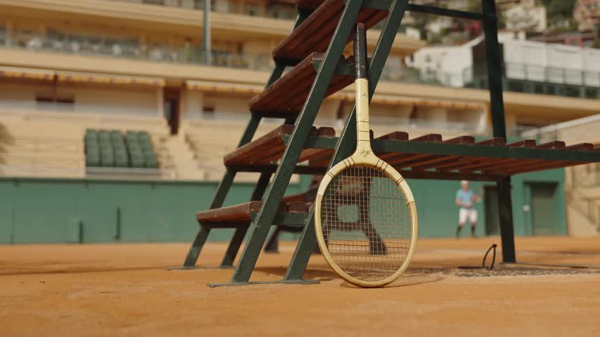 A vintage wooden tennis racket leans against an umpire's chair on a clay court while a player practices in the background