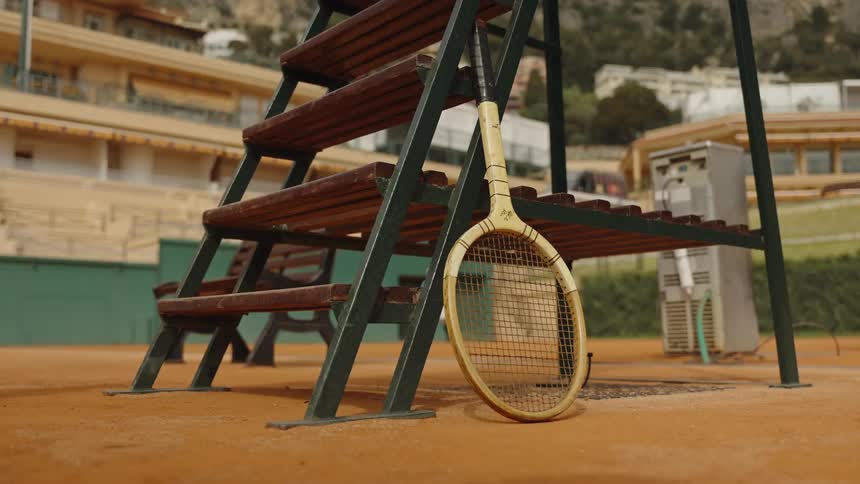 A vintage wooden tennis racket leans against an umpire's chair on a quiet clay court at a stadium