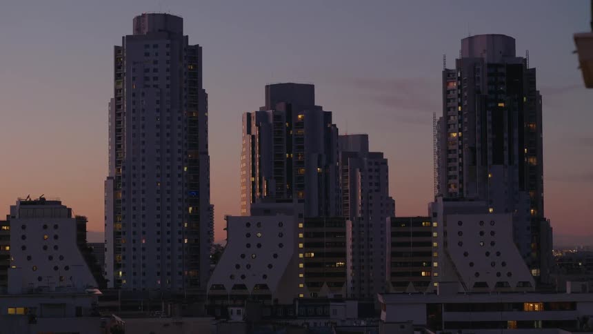 A tranquil view of a modern city skyline with unique apartment buildings illuminated by window lights against a beautiful sunset sky