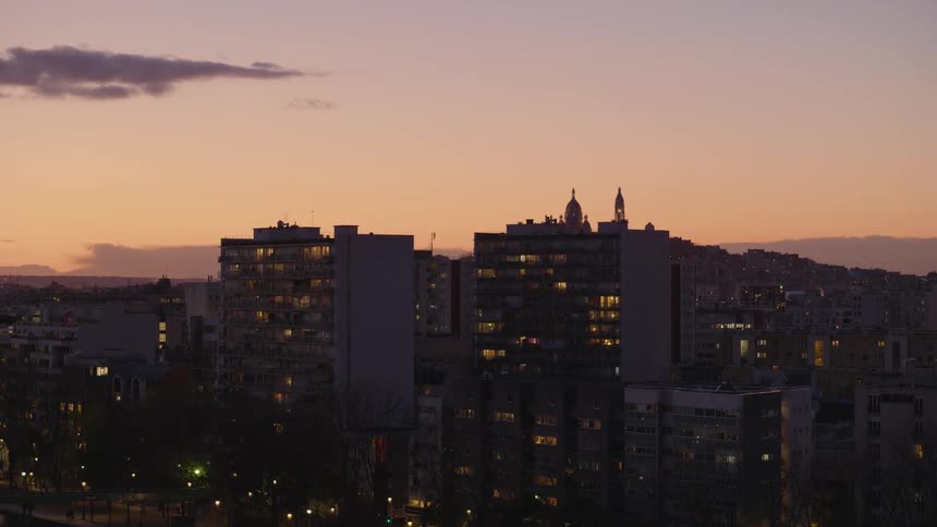 A wide shot of a city skyline at dusk, with lights illuminating the windows of apartment buildings against a warm, colorful sky