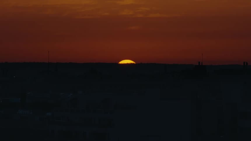 A dramatic timelapse captures the sun sinking below the horizon, painting the sky in deep shades of orange and red over a silhouetted city