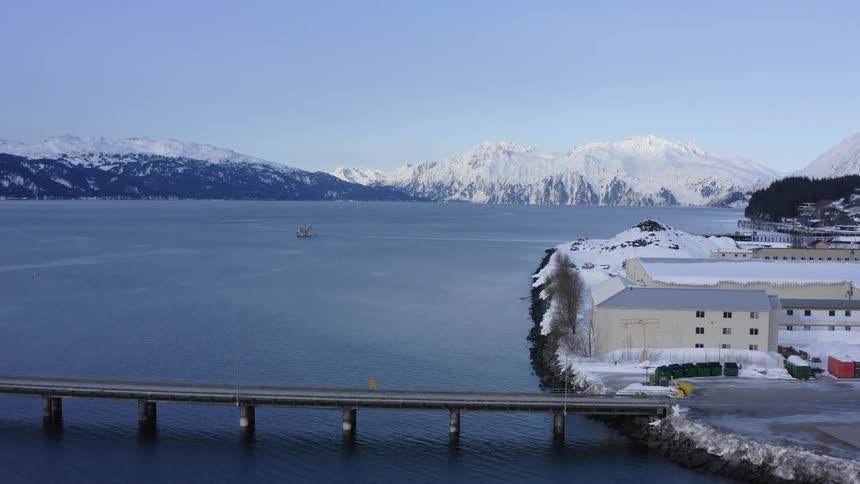 A stunning aerial view captures the serene beauty of a snowy harbor town, complete with a bridge over the water and majestic, snow-covered mountains in the background