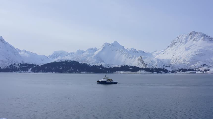 A tugboat navigates a calm bay surrounded by a vast, snow-covered mountain range in winter