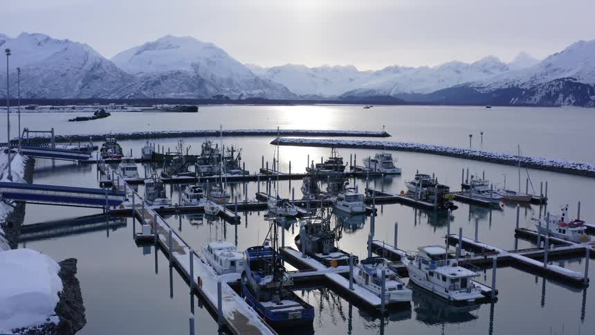 An aerial view of a tranquil, snow-covered harbor with fishing boats docked against a backdrop of majestic mountains
