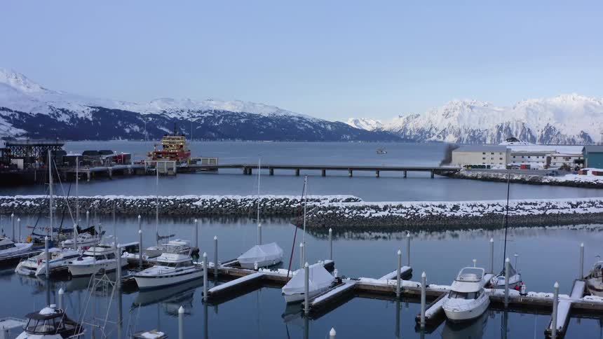Slow aerial flight over the tranquil, snow-covered small boat harbor in Valdez, Alaska, surrounded by towering mountains