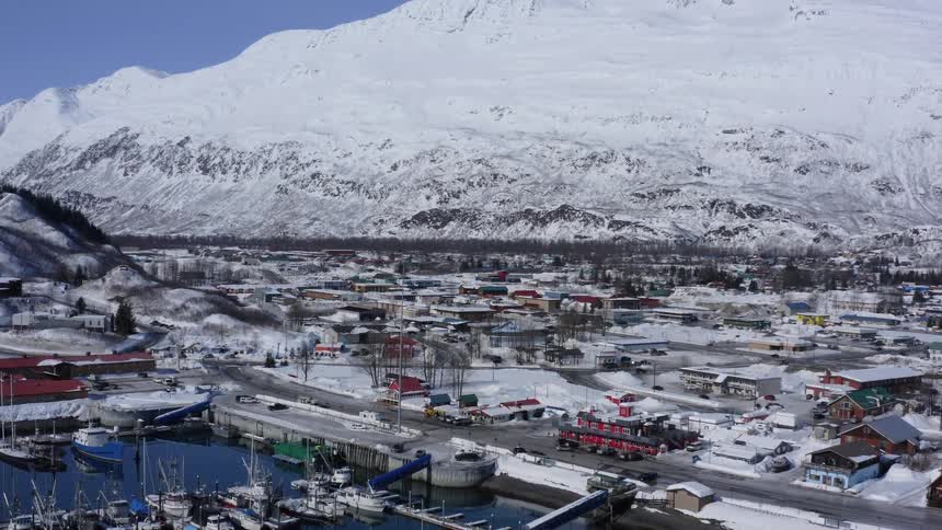An aerial view of the snow-covered coastal town and harbor of Valdez, Alaska, nestled at the foot of the immense Chugach Mountains